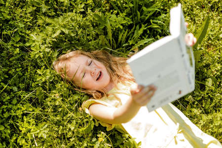 A charming baby is lying on the grass in the garden or park with a book and squinting from the sunの写真素材
