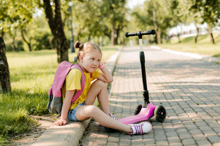 A thoughtful schoolgirl girl is sitting on the sidewalk in the park with a backpack on her shouldersの写真素材
