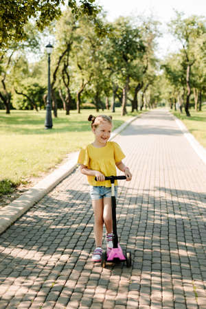 An active little girl rides a scooter on a path in an outdoor park on a summer day. Seasonal childrens active sport. Healthy lifestyle in childhoodの写真素材