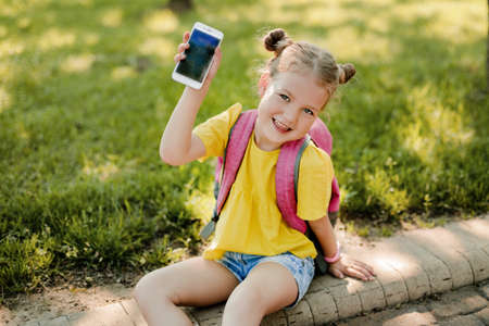 A charming little girl with a backpack is sitting on the sidewalk after school and showing her phone. Back to school, after school, school ageの写真素材