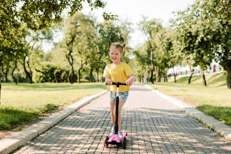 An active little girl rides a scooter on a path in an outdoor park on a summer day. Seasonal childrens active sport. Healthy lifestyle in childhoodの写真素材