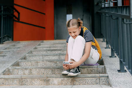 A teenage girl writes messages to friends while sitting on the stairs near the house, on the way to schoolの写真素材