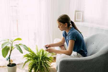A young girl is sitting in a chair and exchanging messages online on her phone. Home recreation,...