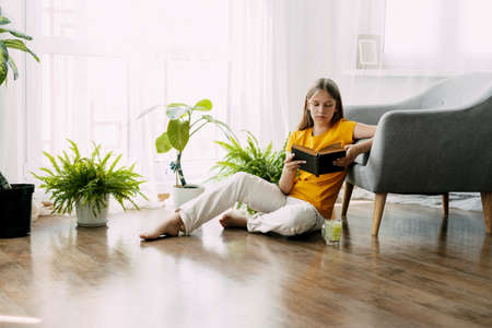 An attractive girl is reading a book, sitting on the floor of the house, having a good time. Rest, relaxationの写真素材