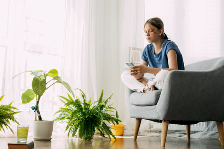 A young girl is sitting in a chair and exchanging messages online on her phone. Home recreation, modern lifestyleの写真素材
