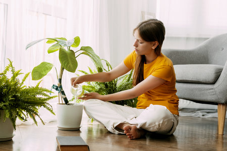 A young happy girl is watering potted plants with water in her small home garden. She is sitting cross-legged on the floorの写真素材