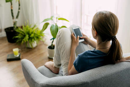A young girl is sitting in a chair and exchanging messages online on her phone. Home recreation,...