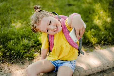 A charming little girl with a backpack is sitting on the sidewalk after school and showing her phone. Back to school, after school, school ageの写真素材