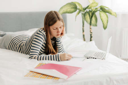 A charming teenage girl is lying on the bed, doing homework with a laptop, making notes in a notebook. The concept of training and education, modern technologiesの写真素材
