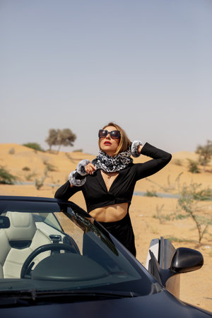 A stylish girl in a black suit and a silk scarf enjoys a ride in a convertible car in the desert, Dubai, UAEの写真素材