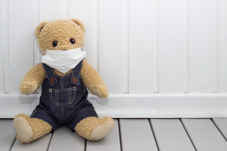 Child's stuffed toy. Brown teddy bear in denim overalls wears a medical disposable mask.の写真素材