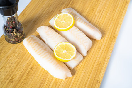 Frozen pieces of cod fish fillets or loins with slices of lemon on cutting board.