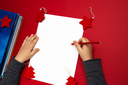Child's hands writing a letter to Santa Clause. Blank sheet of white paper on red background with baubles and stack of fairy books.の写真素材