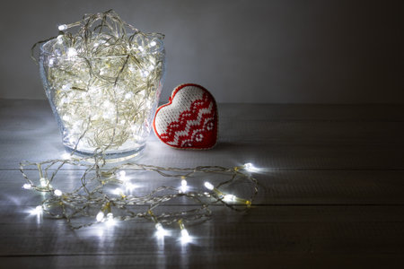 Christmas garland lights in clear glass vase and knitted heart on wooden boards. Copy space.