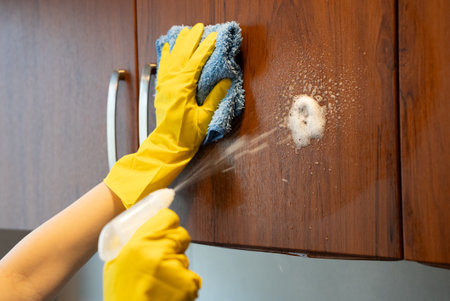 Yellow gloves holding spray bottle and cloth while disinfecting wooden kitchen cabinet. Hygiene and...