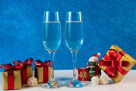 Two champagne glasses surrounded by red and golden Christmas gifts and festive ornaments on a blue background.の写真素材
