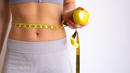 Close-up of slim female waist with measuring tape and apple, symbol of weight loss and body goals.の写真素材