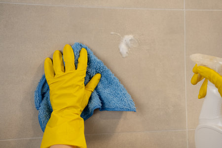 Close-up of hand in yellow glove using spray cleaner and blue microfiber cloth on tiled kitchen wall, concept of sanitation and spotless hygiene.の写真素材
