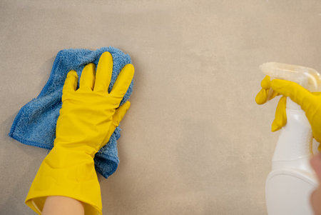 Close-up of female hand wearing yellow gloves with cleaning cloth against neutral background....
