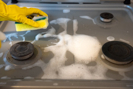 Close-up of gloved hand scrubbing dirty gas stove burner with sponge and detergent foam, concept of kitchen hygiene and disinfection.の写真素材