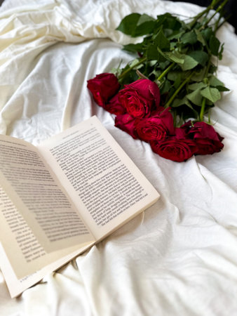Open book with red roses on white bedding in soft light. Cozy romantic flat lay for reading, love and slow lifestyle concepts. Open book with red roses on white beddingの写真素材