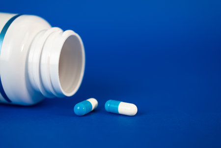 Blue and white capsules lying near an open white bottle on a blue background. Pharmaceutical,...