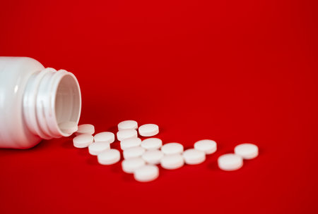 White pills scattered from open bottle on red background, representing healthcare, treatment, and...