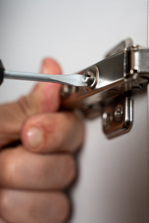 Close-up of a hand using a screwdriver to repair or adjust a metal cabinet hinge. DIY, service,...
