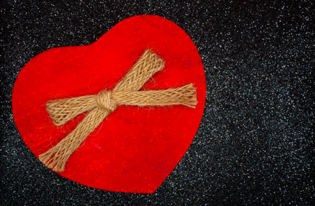 Single red heart shaped gift tied with rustic ribbon on black glitter surface. Romantic holiday...