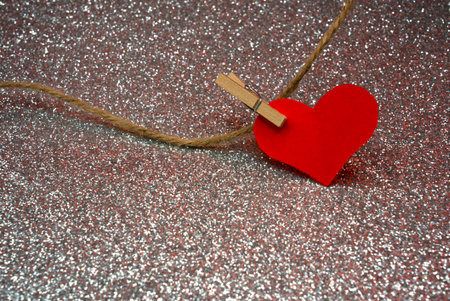 Small red heart decoration hanging on string over silver glitter surface. Minimal romantic...