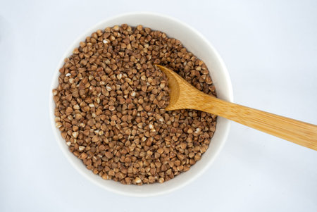 Top view of raw buckwheat groats in a white ceramic bowl with wooden spoon on a neutral background. Healthy food concept, clean minimal still life for nutrition and diet themes.の写真素材