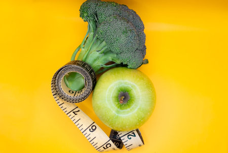 Fresh green broccoli and apple with a measuring tape on a vibrant yellow background, representing...