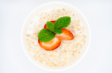 Overhead view of a white bowl filled with creamy oatmeal, topped with fresh strawberry slices and...