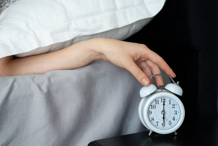 Close-up of a woman's hand reaching for an alarm clock on a nightstand beside her bed, with her body...