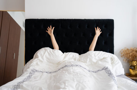 A woman lies in bed with her hands raised, surrounded by white bedding and a black headboard, in a...