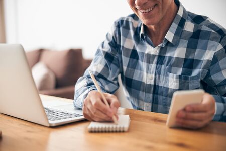 Cheerful adult man sitting at his workplace and making notes during online conference, enjoying his working process at homeの写真素材