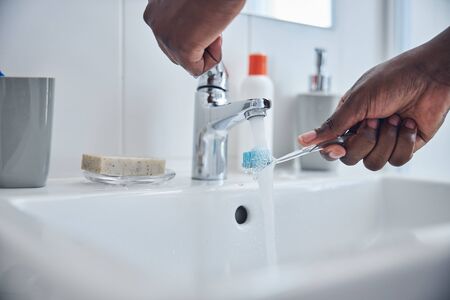 Focused photo on dark-skinned male person that standing near sink and putting brush under the water before cleaning teethの写真素材