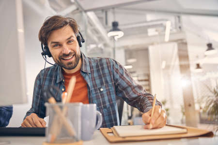 Cheerful young man writing in spiral notebook at work
