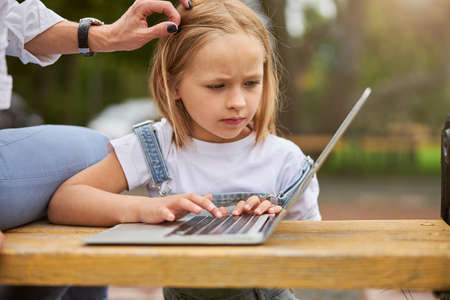 Portrait of charming focused girl at the lesson on laptop in the park with mom