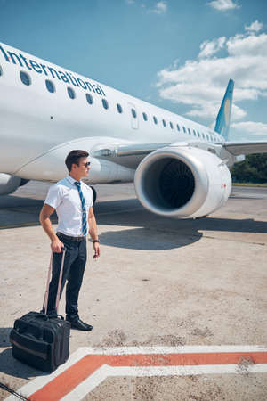 Handsome male pilot with travel suitcase standing in airfieldの写真素材