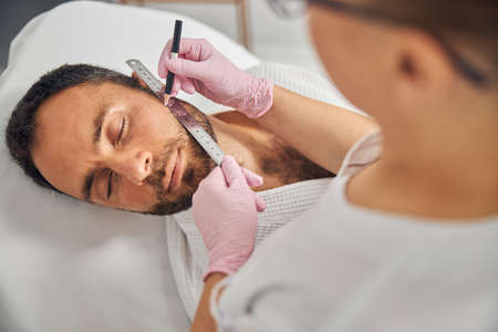 Female esthetician hands drawing beard cheek line on man skin