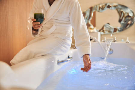 Young man in bathrobe sitting on edge of bathtubの写真素材