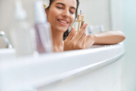 Smiling young woman holding bottle of face serum while taking bathの写真素材
