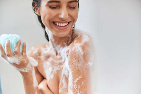 Cheerful young woman with foam on her body holding bath loofah