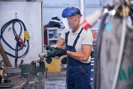 Bearded mechanic in protective mask using grinder in garageの写真素材