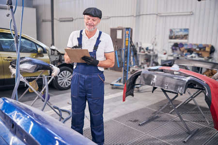 Joyful auto mechanic writing on clipboard at car repair service station