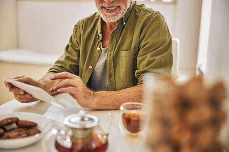Positive senior citizen having tea and clicking on his tabletの写真素材