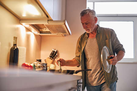 Senior gentleman lifting the lid off frying pan on cookerの写真素材