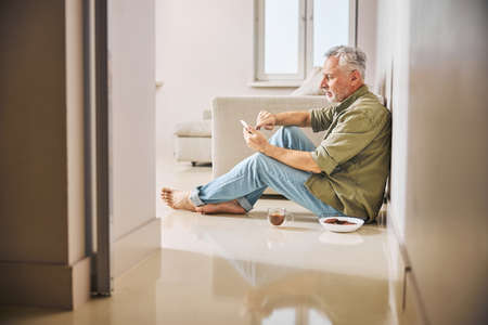 Senior man looking at his phone while sitting on floorの写真素材