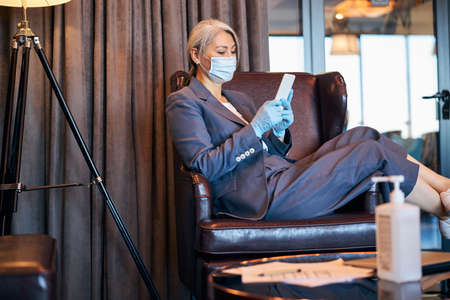 Elegant businesswoman in medical mask using mobile phoneの写真素材